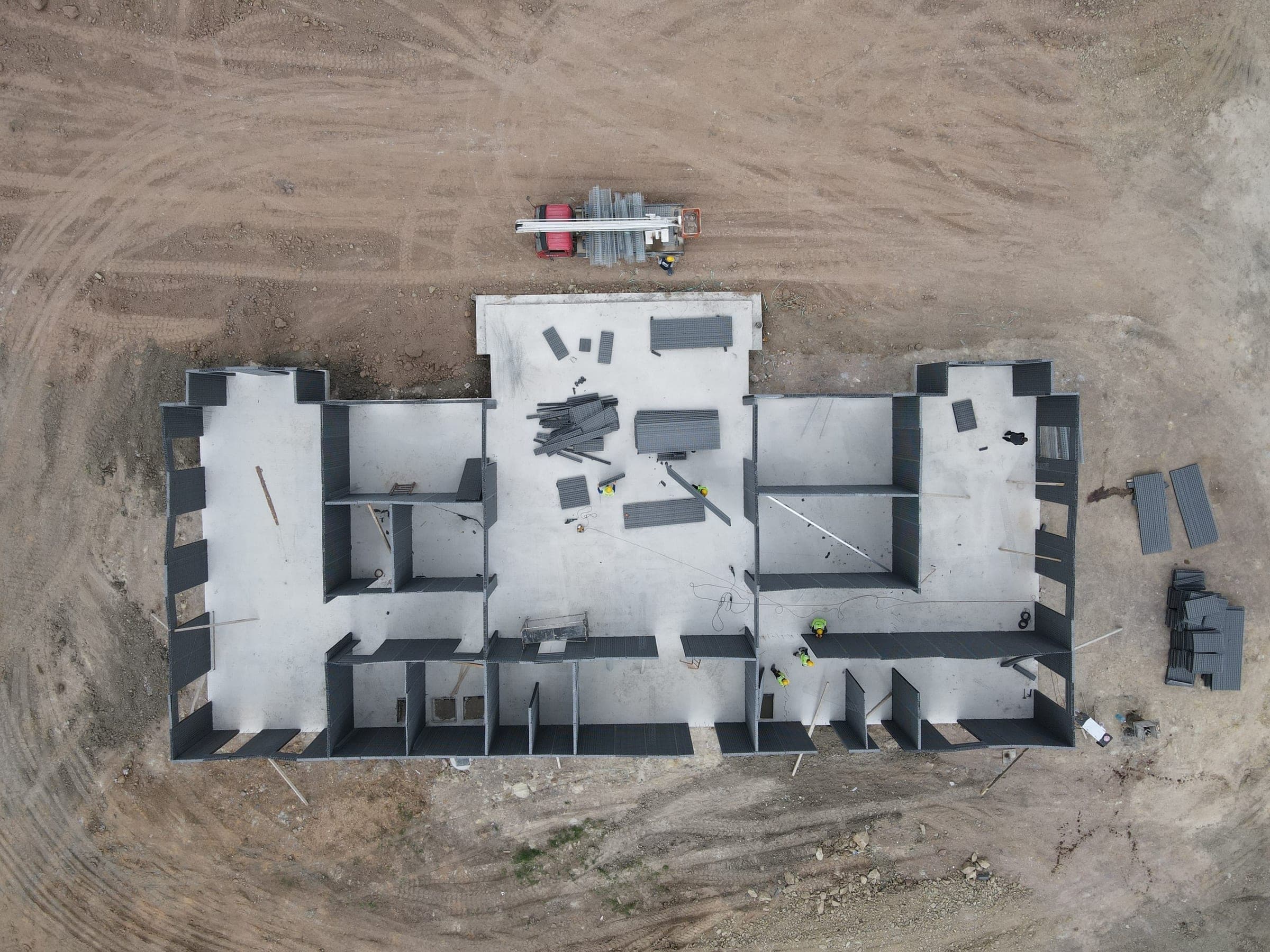 Aerial view of a foundation slab with the first wall panels in place and a truck delivering more panels.