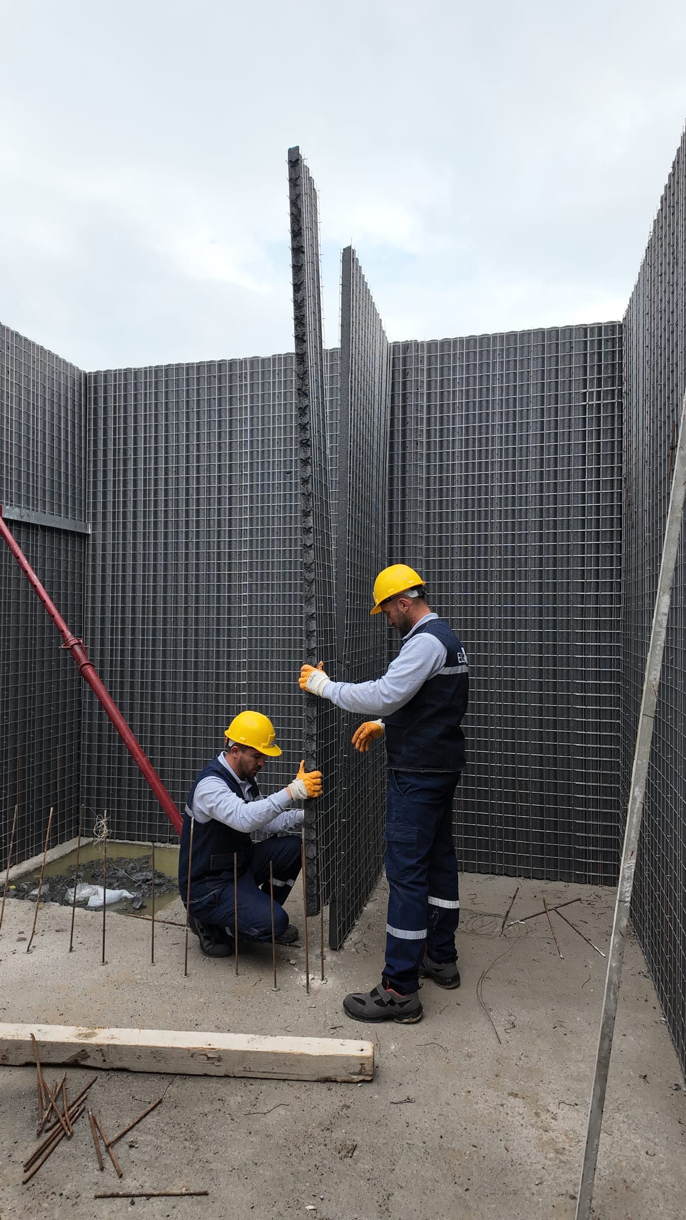 Two workers in hard hats setting a steel-mesh panel upright in a narrow space, braced with a steel prop.