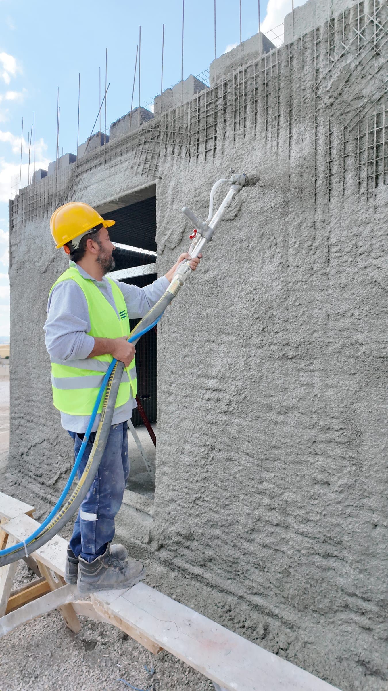 A worker spraying shotcrete onto a steel-mesh panel wall; the fresh surface is rough and grey.